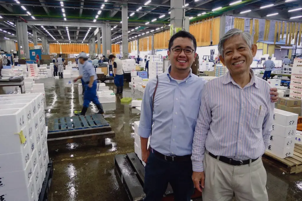 A eye-level medium portrait shot shows two generations of founders posing together at a busy wholesale seafood port.