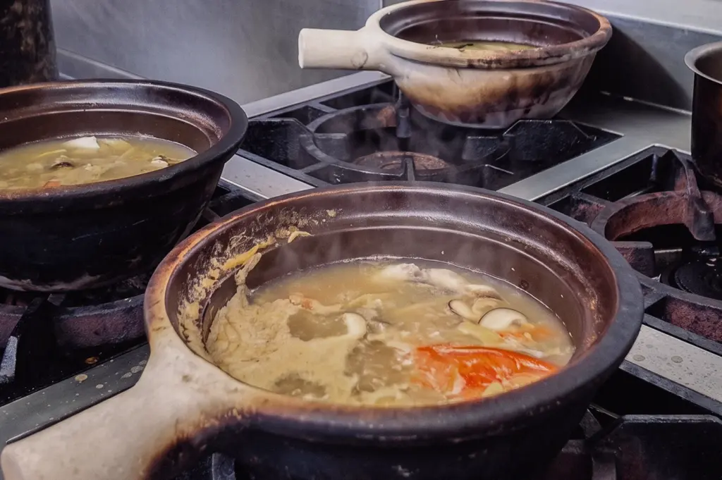 A high-angle close-up shot captures claypots simmering with collagen fish soup over gas flames on a commercial stove burner.