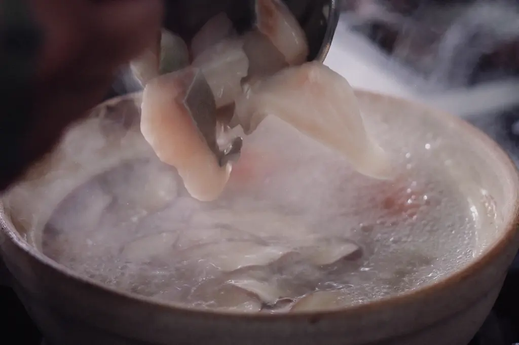 A close-up action shot showing fresh pomfret fish slices being added into a boiling claypot of simmering clear broth.
