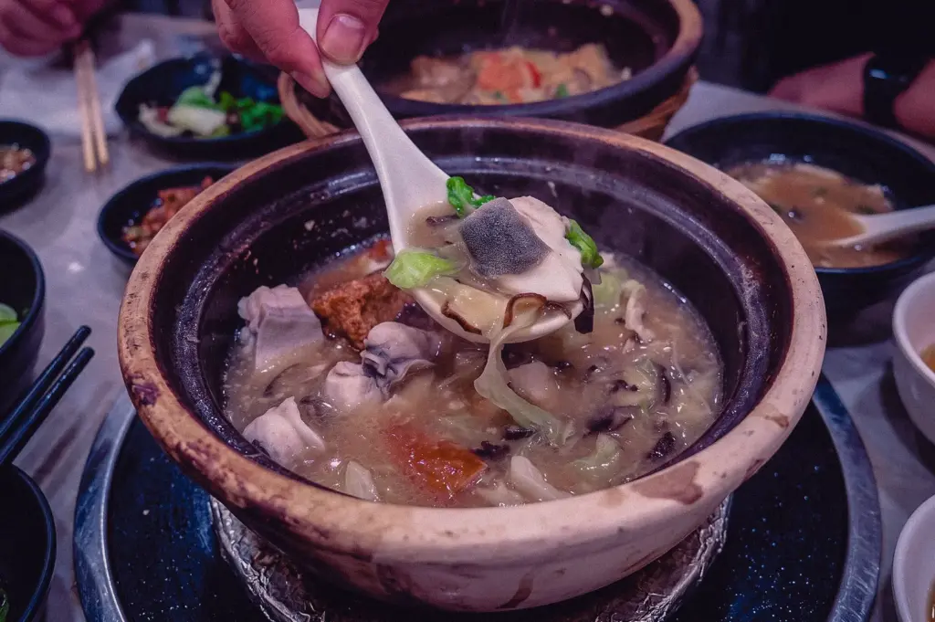 High-angle close-up shot capturing a white spoon scooping a tender slice of pomfret fish and leafy greens from a claypot of collagen-rich soup.