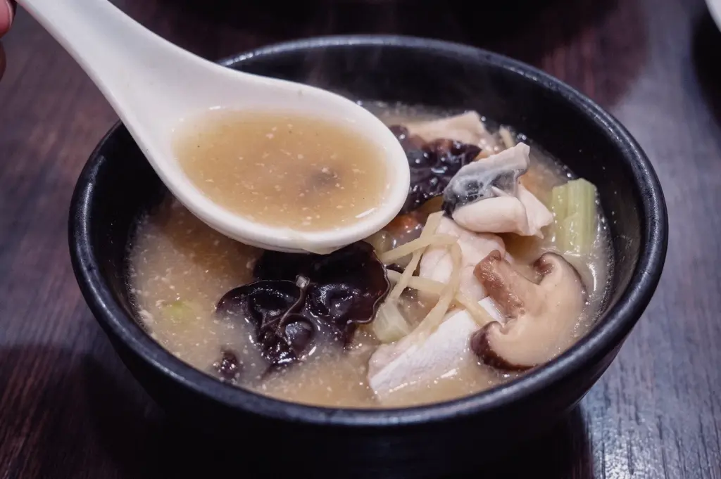 High-angle close-up shot showing a white ceramic spoon filled with rich collagen broth served over a black bowl of pomfret slices, wood ear mushrooms, and sliced mushrooms.