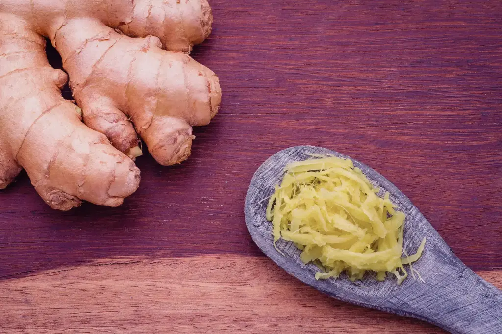 A high-angle close-up shot shows finely shredded julienned ginger on a wooden spoon alongside a fresh ginger root on a dark wooden surface.