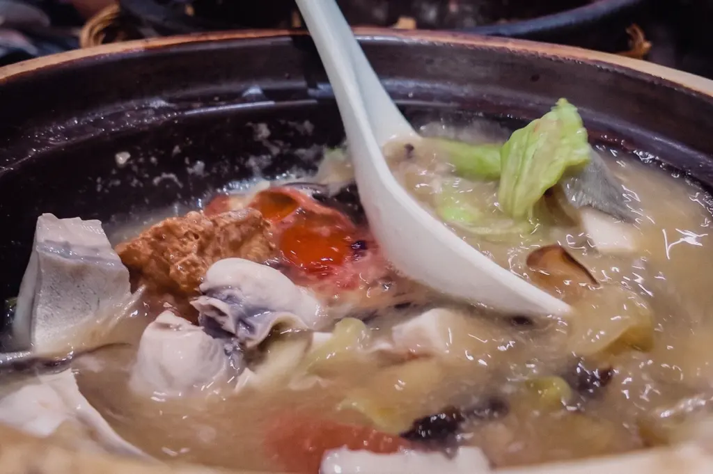 A high-angle close-up shot shows a spoon scooping rich gelatinous broth from a claypot containing pomfret slices and fried tofu.