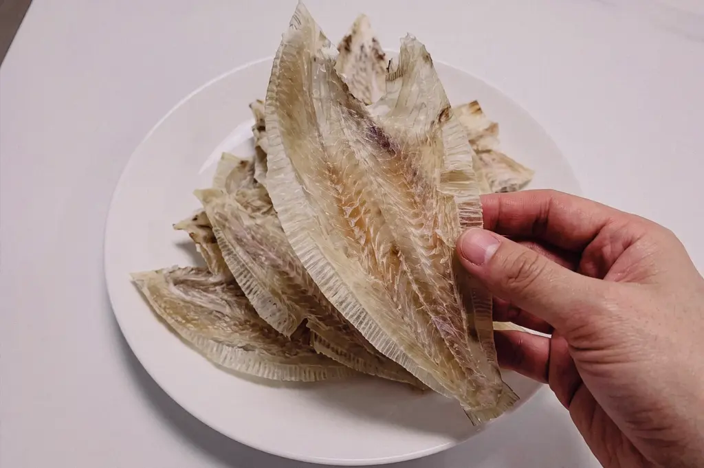Top-down close-up shot of a hand holding a dried tee poh sole fish fillet over a white plate to illustrate the natural umami base.