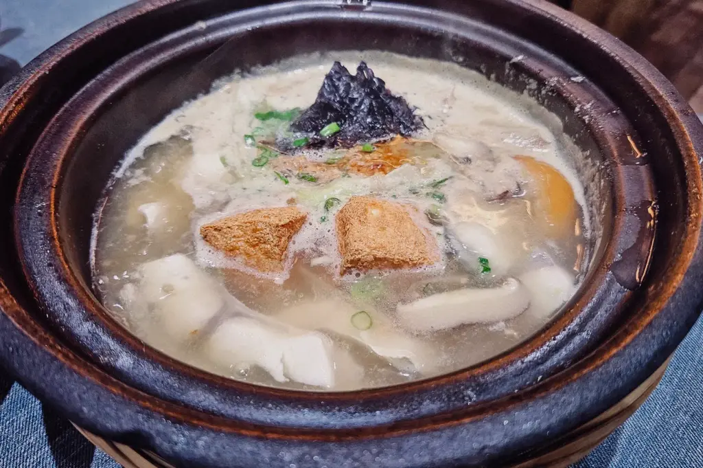 A top-down overhead flat lay shot displays a piping-hot claypot of milky ivory fish soup topped with fried tofu and seaweed.