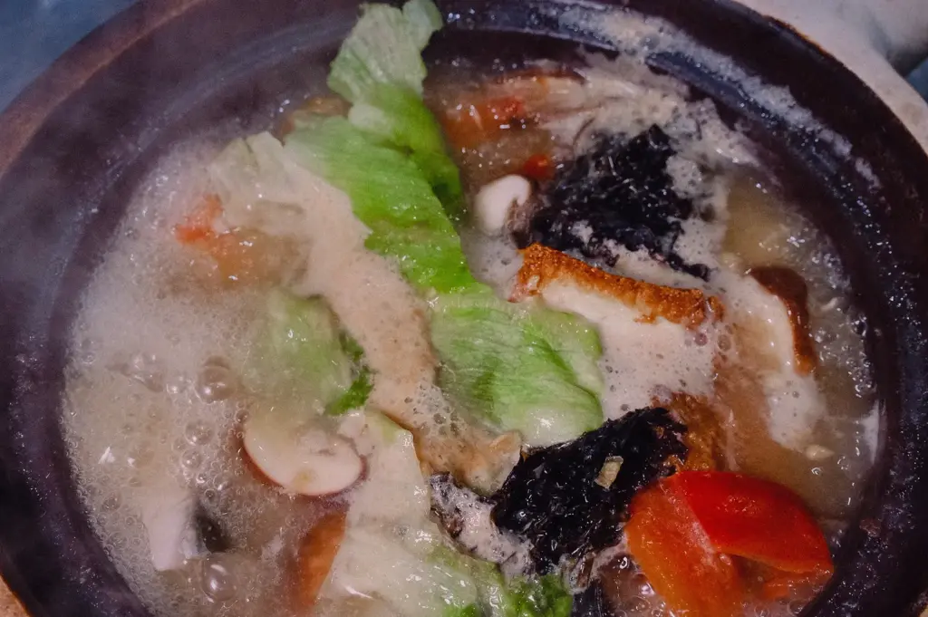 A top-down overhead shot shows a claypot of Grandpa's Pomfret Soup bubbling gently over residual heat with fresh lettuce, seaweed, and tomatoes.