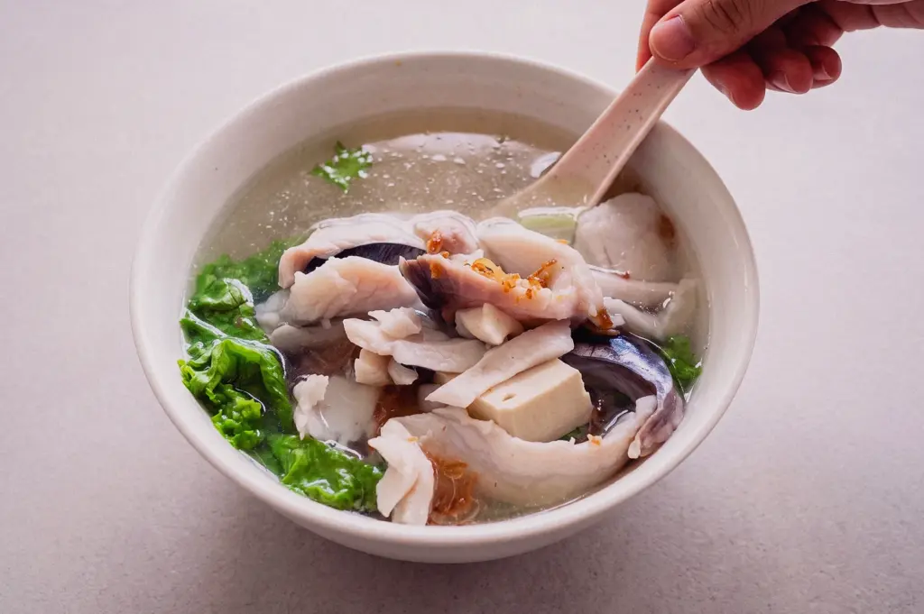 A high-angle close-up shot captures a spoon scooping tender fish slices from a bowl of clean Teochew fish soup.