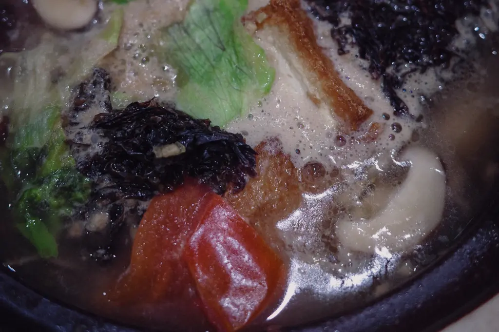 Macro top-down shot detailing the rich, simmering ivory fish broth garnished with seaweed, tomatoes, and leafy vegetables inside a traditional claypot.