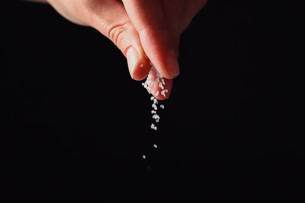 A close-up action shot captures a person's fingers pinching coarse sea salt crystals against a dark background.