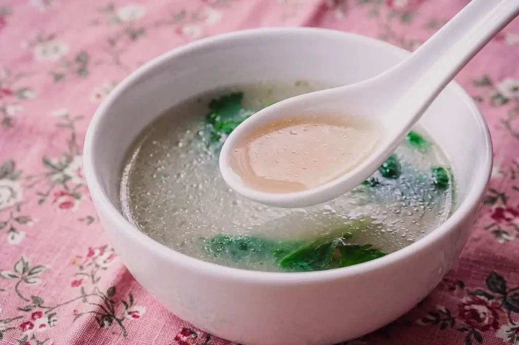 A high-angle three-quarter shot features a white ceramic spoon holding clear, golden fish soup broth over a bowl garnished with leafy greens.