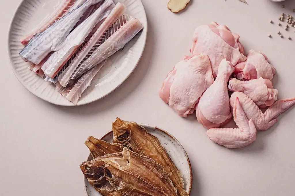 High-angle flat lay view displaying the key soup broth ingredients arranged on a light surface, including raw batang fish bones on a ceramic plate, fresh raw chicken cuts, and dried tee poh sole fillets.