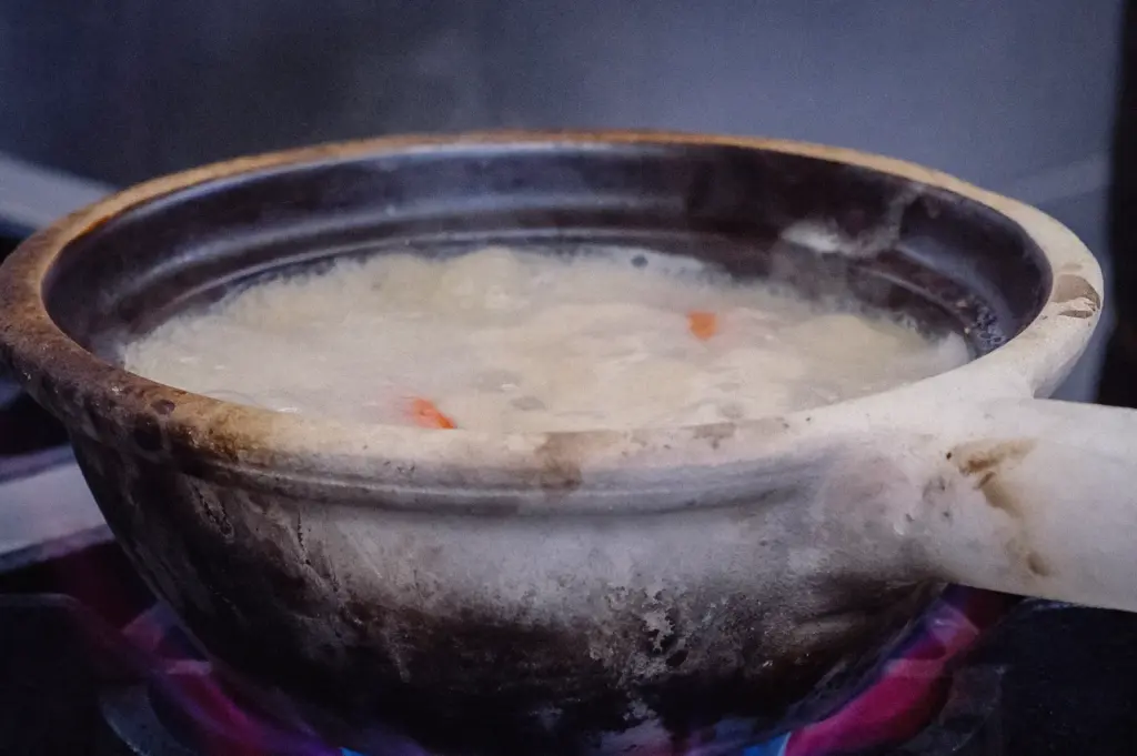 A low side-angle close-up shot captures milky white collagen fish broth simmering inside a traditional claypot over open gas stove flames.