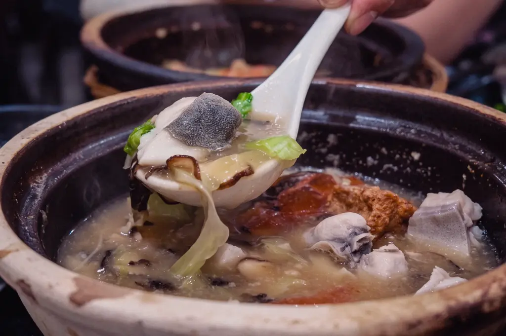 A close-up action shot features a white spoon lifting a thick pomfret fish slice with cabbage and mushrooms from a bubbling claypot of Grandpa's Pomfret Soup.