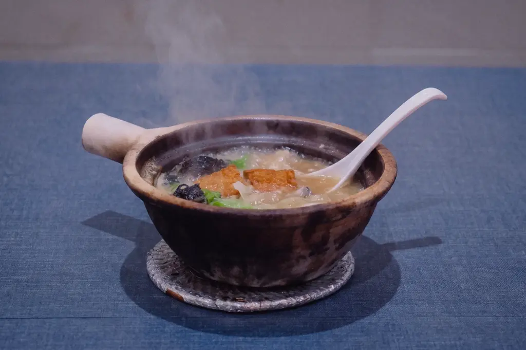 A eye-level medium shot displays a steaming claypot of Grandpa's Pomfret Soup topped with tofu and greens resting on a woven coaster over a blue table.