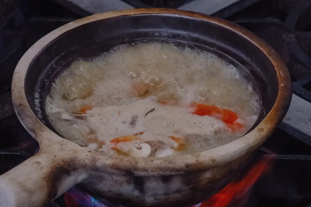 A high-angle close-up shot highlights a claypot gently bubbling over an open stove flame with collagen broth, tomatoes, and ingredients.