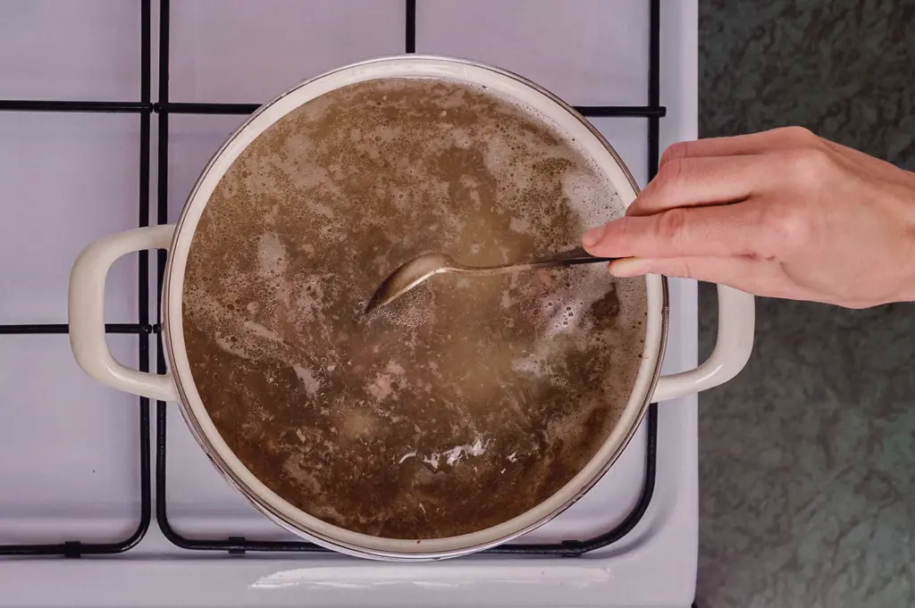 A top-down overhead flat lay shot features a hand stirring a large pot of simmering clear fish broth on a white stovetop.