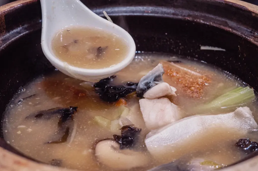 An eye-level close-up shot featuring a spoonful of rich collagen broth scooped from a claypot filled with tender pomfret fish, mushrooms, and ingredients.