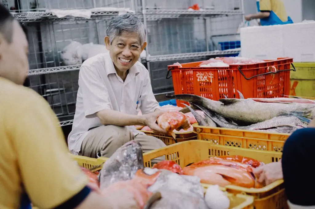 Eye-level medium shot featuring an elderly fishmonger smiling while inspecting fresh red snapper and whole fish in plastic crates at Jurong Fishery Port.