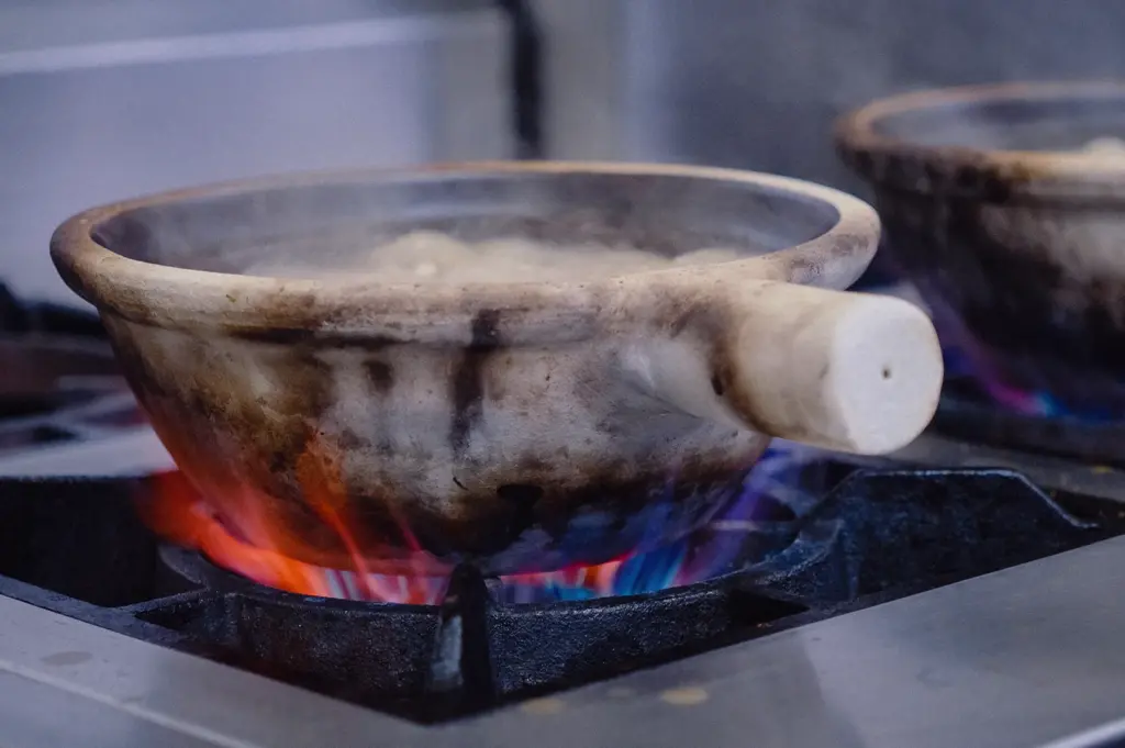 Low-angle close-up shot capturing a claypot simmering over a bright blue stove flame in a commercial kitchen.