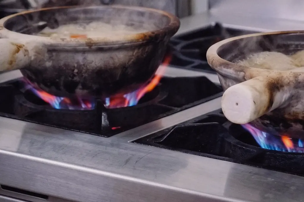 A low-angle three-quarter shot shows claypots simmering over controlled gas stove flames to extract collagen for clean fish broth.