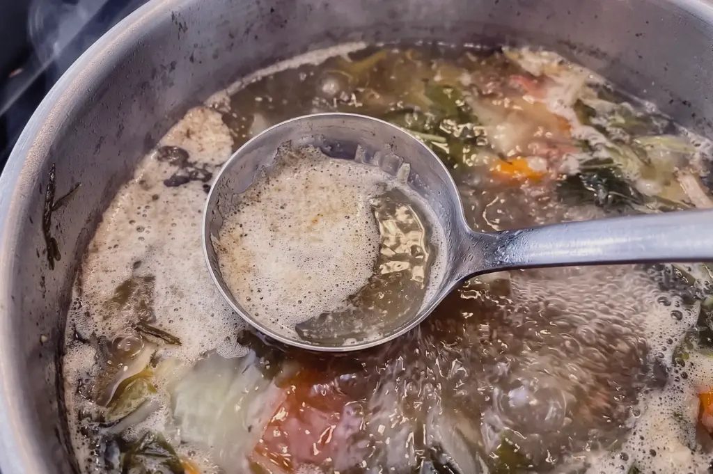 Top-down close-up shot showing a metal ladle skimming floating scum and foam from a simmering pot of fish stock.