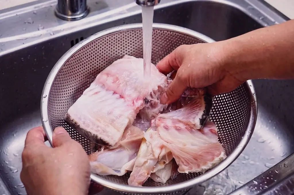 A high-angle close-up action shot shows fresh fish bones being rinsed under running tap water in a stainless steel colander to remove blood and prevent bitter off-notes.