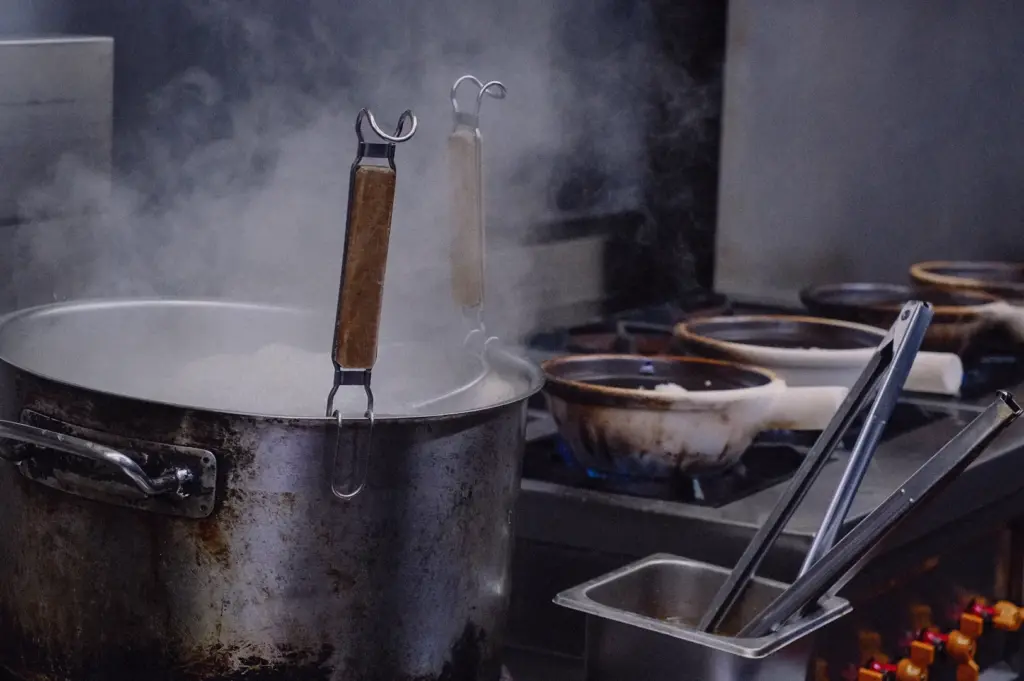 Eye-level medium shot showing a large stainless steel stockpot steaming on a commercial stove next to claypots and cooking utensils in the Hup Lok kitchen.