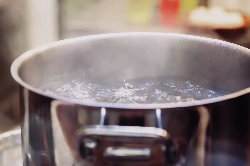 A side-angle close-up shot features fish soup broth rapidly boiling and steam rising inside a stainless steel stockpot on a gas stove.