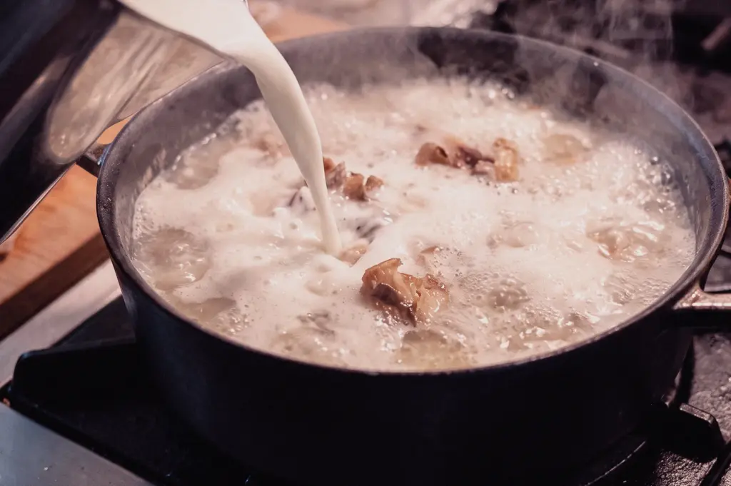 A side-angle close-up shot shows evaporated milk being poured into a simmering stockpot of fish soup over high heat.