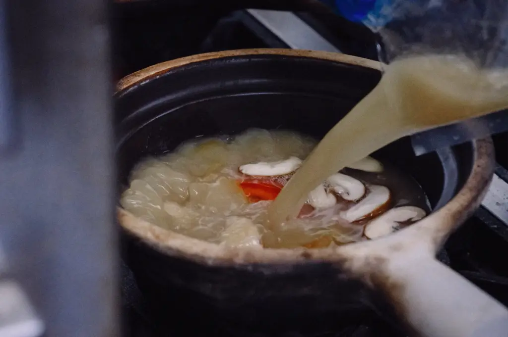 Close-up action shot of rich collagen fish broth being poured into a claypot with tomatoes and mushrooms.