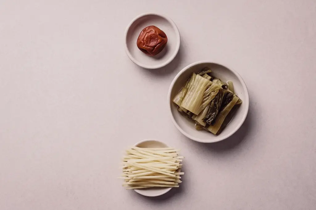 Overhead flat lay shot showcasing three small ceramic bowls containing traditional Teochew soup condiments: a single sng buay sour plum, sliced kiam chye salted mustard greens, and fine julienned ginger.