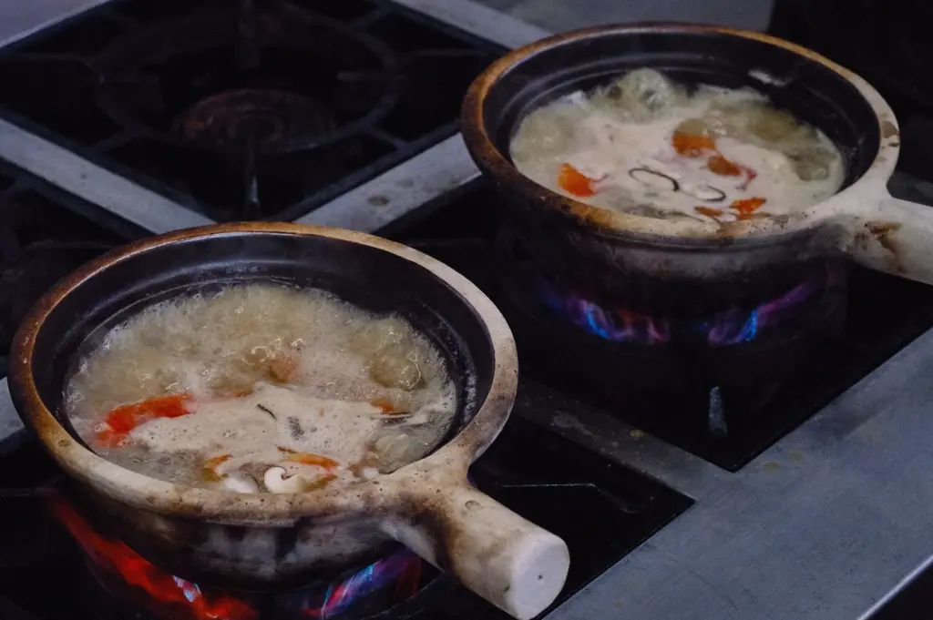 A high-angle three-quarter view shows two claypots of fish soup bubbling simultaneously on commercial gas stove burners.