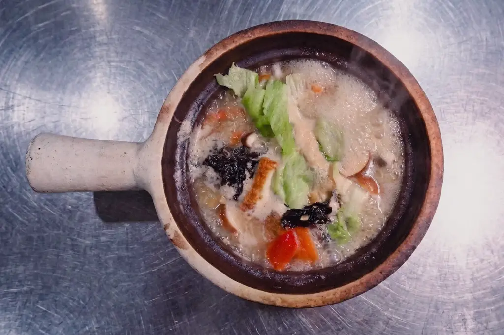 A directly overhead flat lay shot displays a hot claypot of pomfret fish soup resting on a stainless steel tabletop, showing cabbage leaves, tomatoes, and rich broth.