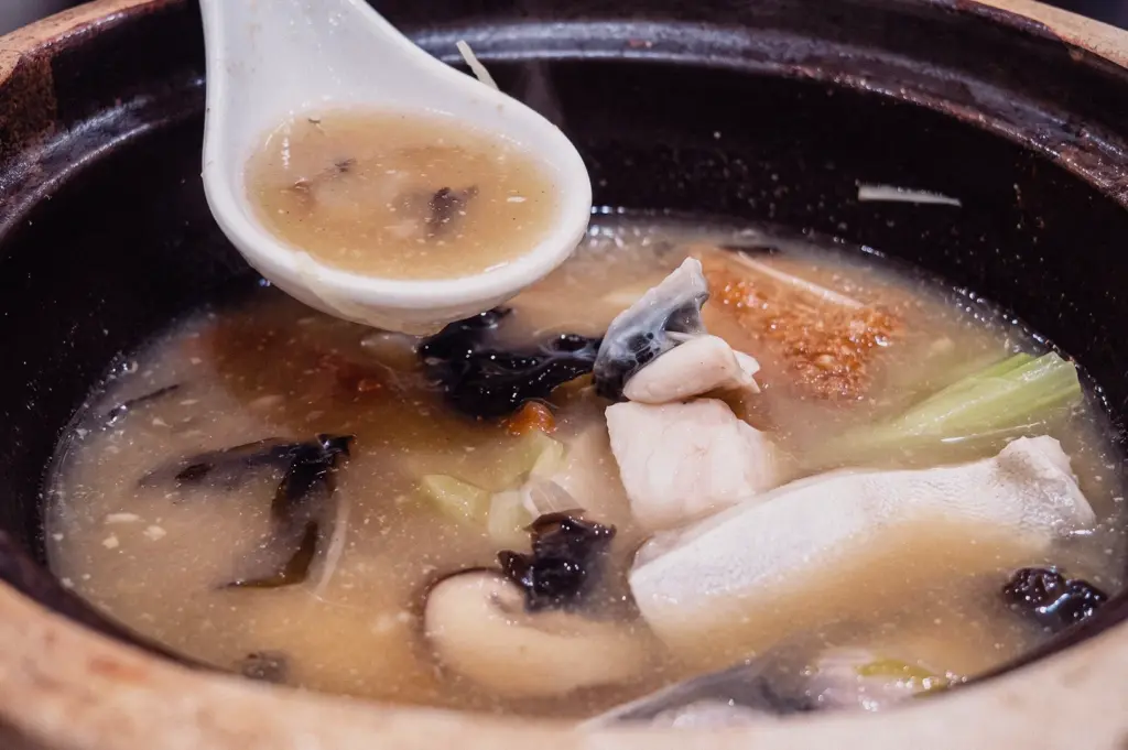 An eye-level close-up shot features a white ceramic spoon scooping rich, collagen-packed fish broth from a hot claypot containing pomfret slices and wood ear mushrooms.