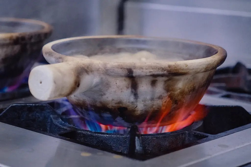 A low-angle close-up shot focuses on a traditional claypot sitting over blue and orange gas flames as the collagen fish broth comes to a steady boil.