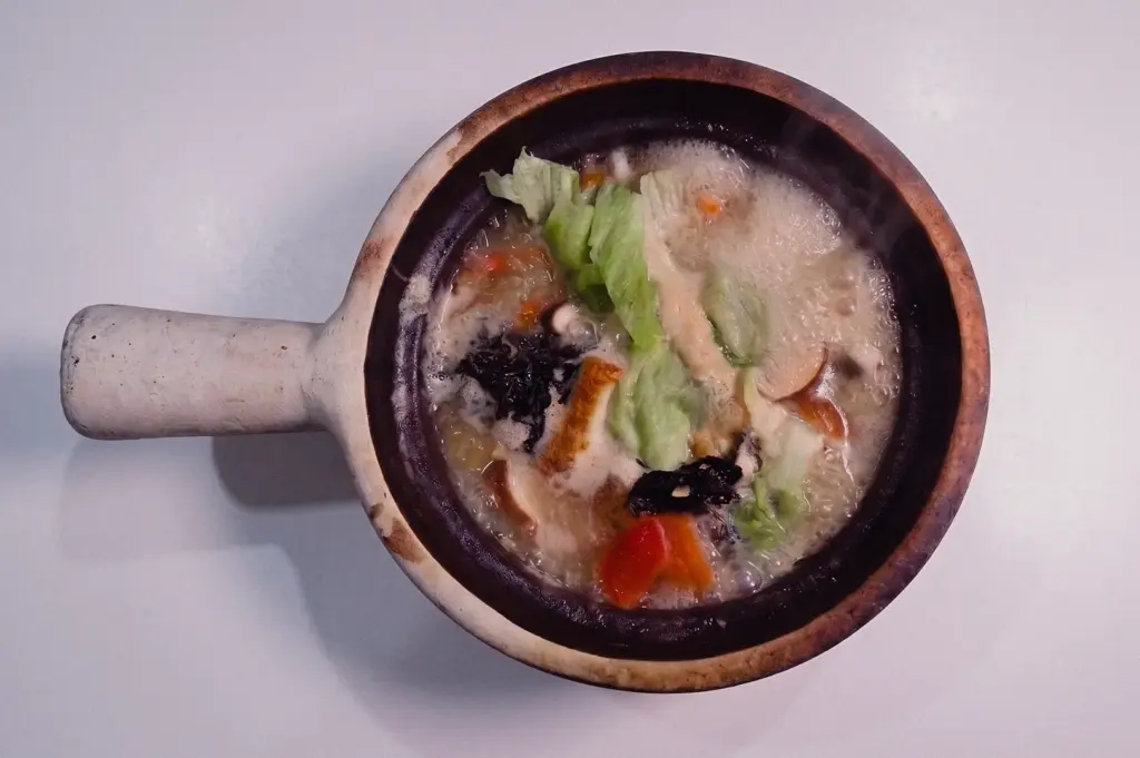 Top-down shot featuring a piping hot claypot of pomfret fish soup garnished with fresh lettuce, seaweed, and tomatoes on a white tabletop.