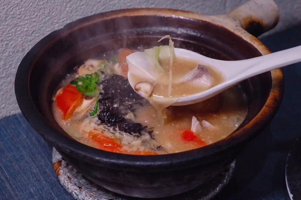 A high-angle close-up shot captures a white ceramic spoon lifting a tender pomfret fish slice with julienned ginger from a steaming claypot of rich, heavy fish soup.