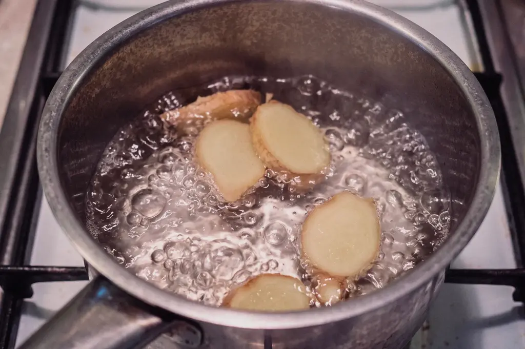 A high-angle close-up shot features fresh ginger slices simmering in boiling water inside a stainless steel saucepan on a gas stove top.