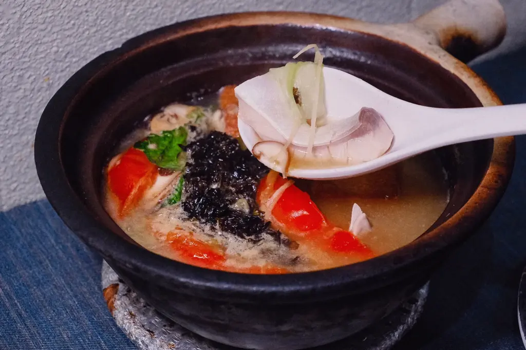 A high-angle close-up shot features a soup spoon lifting a tender pomfret fish slice with julienned ginger above a steaming claypot of Grandpa's Pomfret Soup.