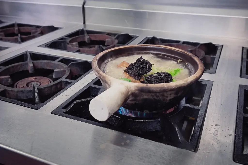 A wide three-quarter angle shot shows a claypot filled with simmering pomfret fish soup and topped with seaweed on a commercial kitchen burner.