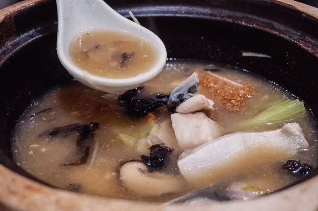 An eye-level close-up shot features a white spoon lifting rich, collagen-packed fish broth from a claypot containing pomfret slices and mushrooms.