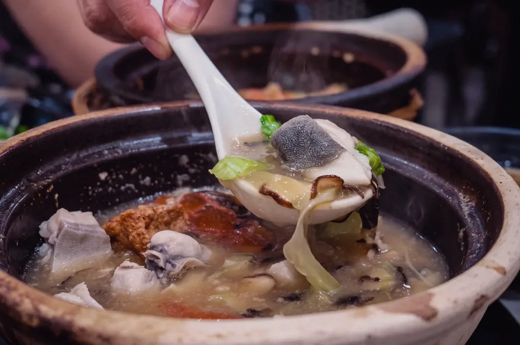 A eye-level close-up action shot shows a hand holding a white spoon lifting a slice of fresh pomfret fish skin-side up over a claypot of steaming broth.