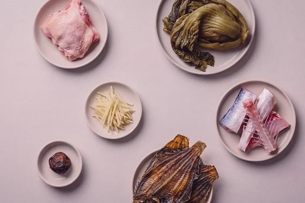 A top-down overhead flat lay shot showcases soup ingredients arranged on individual plates, including raw chicken, salted mustard, julienned ginger, batang fish bones, dried sole fillet (tee poh), and sour plum.