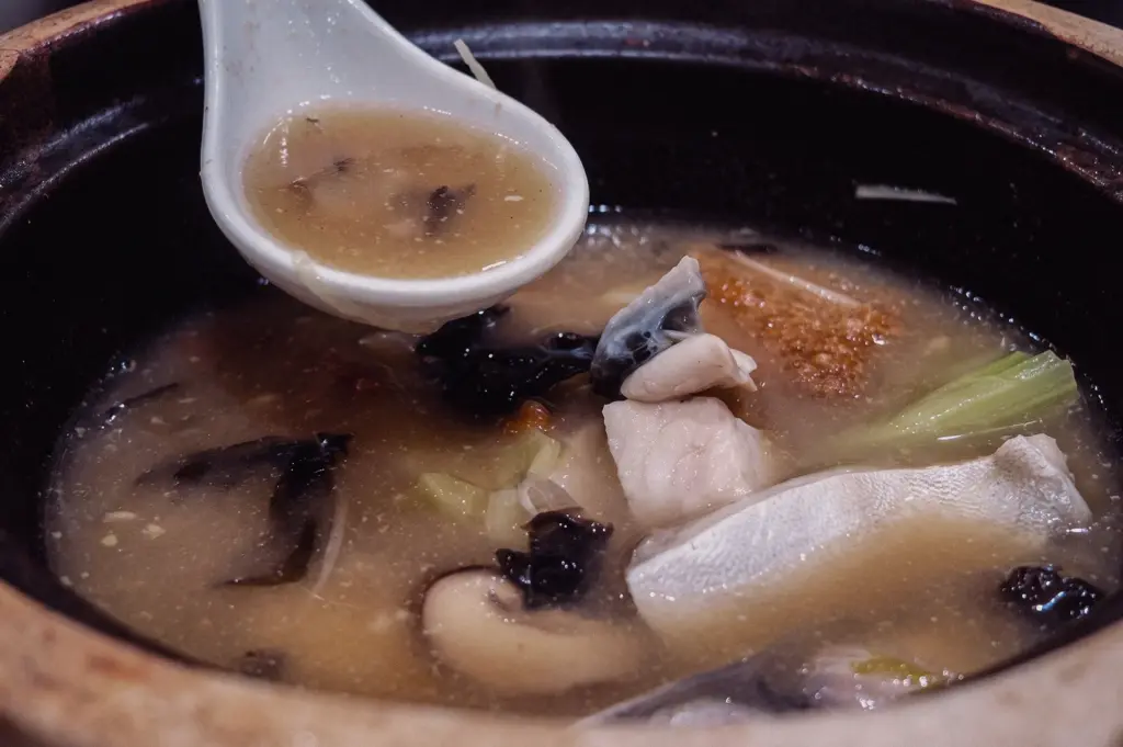 High-angle close-up shot focusing on fresh pomfret fillets, mushrooms, and seaweeds submerged in collagen-rich broth inside a warm claypot dish.