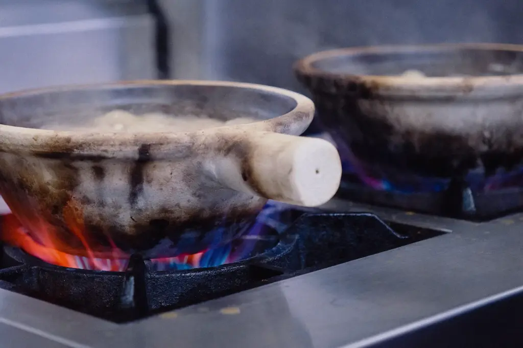 A low-angle close-up shot displays claypots resting over gas stove flames in a commercial kitchen as fish broth boils.