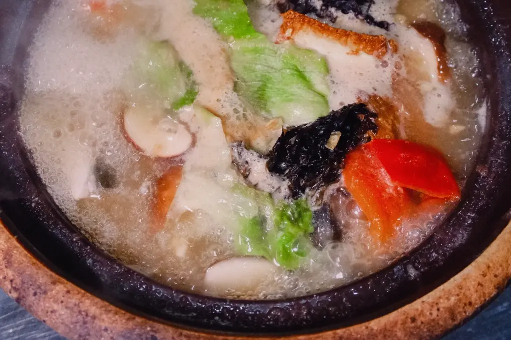 A top-down overhead shot highlights a bubbling claypot of milky white collagen fish soup loaded with tomatoes, greens, mushrooms, and fried tofu.