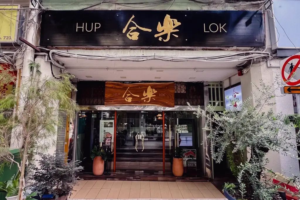 Wide straight-on exterior shot of the Hup Lok storefront on Havelock Road with wooden signboards and potted green plants.