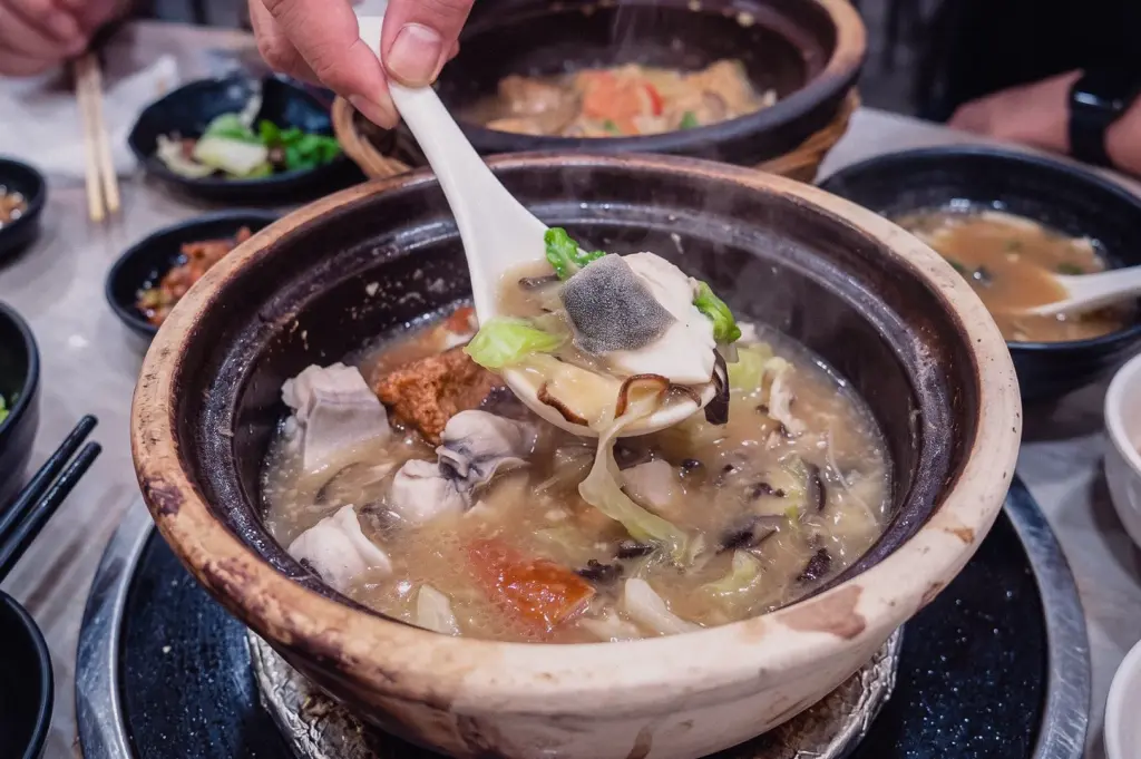 A high-angle close-up action shot shows a spoon lifting tender pomfret fish slices and cabbage from a steaming claypot of Grandpa's Pomfret Soup at a restaurant table.