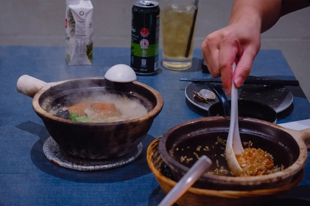 An eye-level medium action shot shows a diner using a white spoon to mix rice in a claypot next to a steaming bowl of Grandpa's Pomfret Soup on a dining table at Hup Lok.