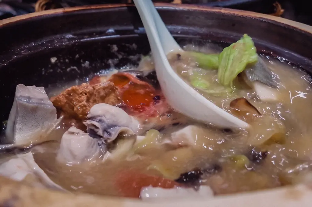 A close-up three-quarter shot captures a white spoon resting inside a hot claypot of Grandpa's Pomfret Soup enriched with fish slices, vegetables, and ginger.